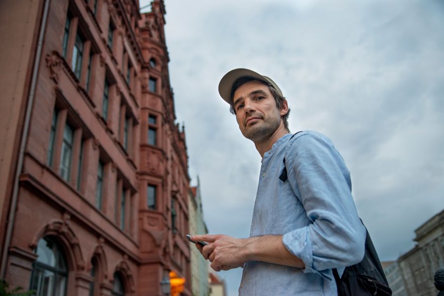 Casually dressed man using smartphone in Berlin city street with historic architecture. — Developers · Qatar: Golden hour, fully online — photo backups upload before dinner.