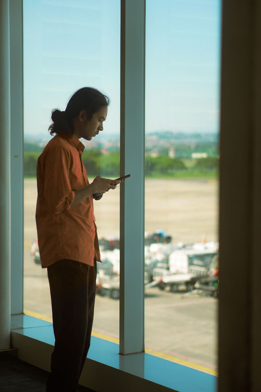 Man in orange shirt using a smartphone while standing at an airport window, overlooking the runway. — Developers · Côte d'Ivoire: The museum queue skips itself: tickets bought online, straight to the door.