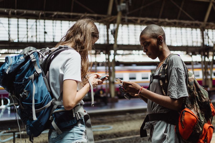 Two adventurous backpackers using smartphones while waiting at a bustling train station. — Developers · Lithuania: Market mornings: look up the dish, translate the menu, pay by phone.